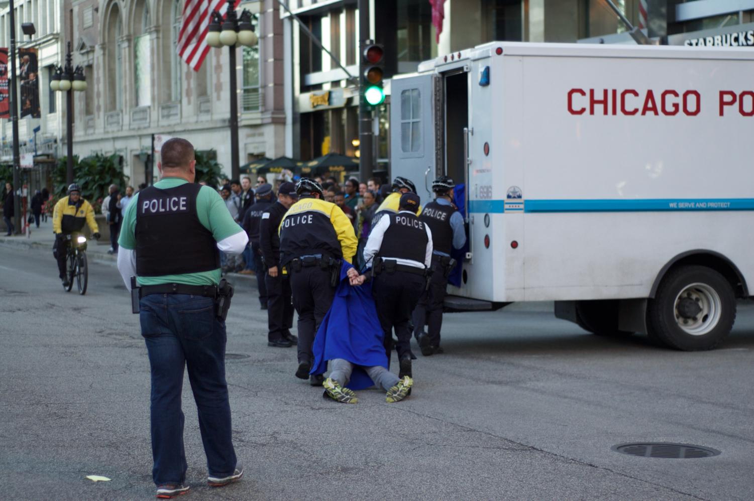 Chicago police carry a limp protestor in a cap and gown into a police van.