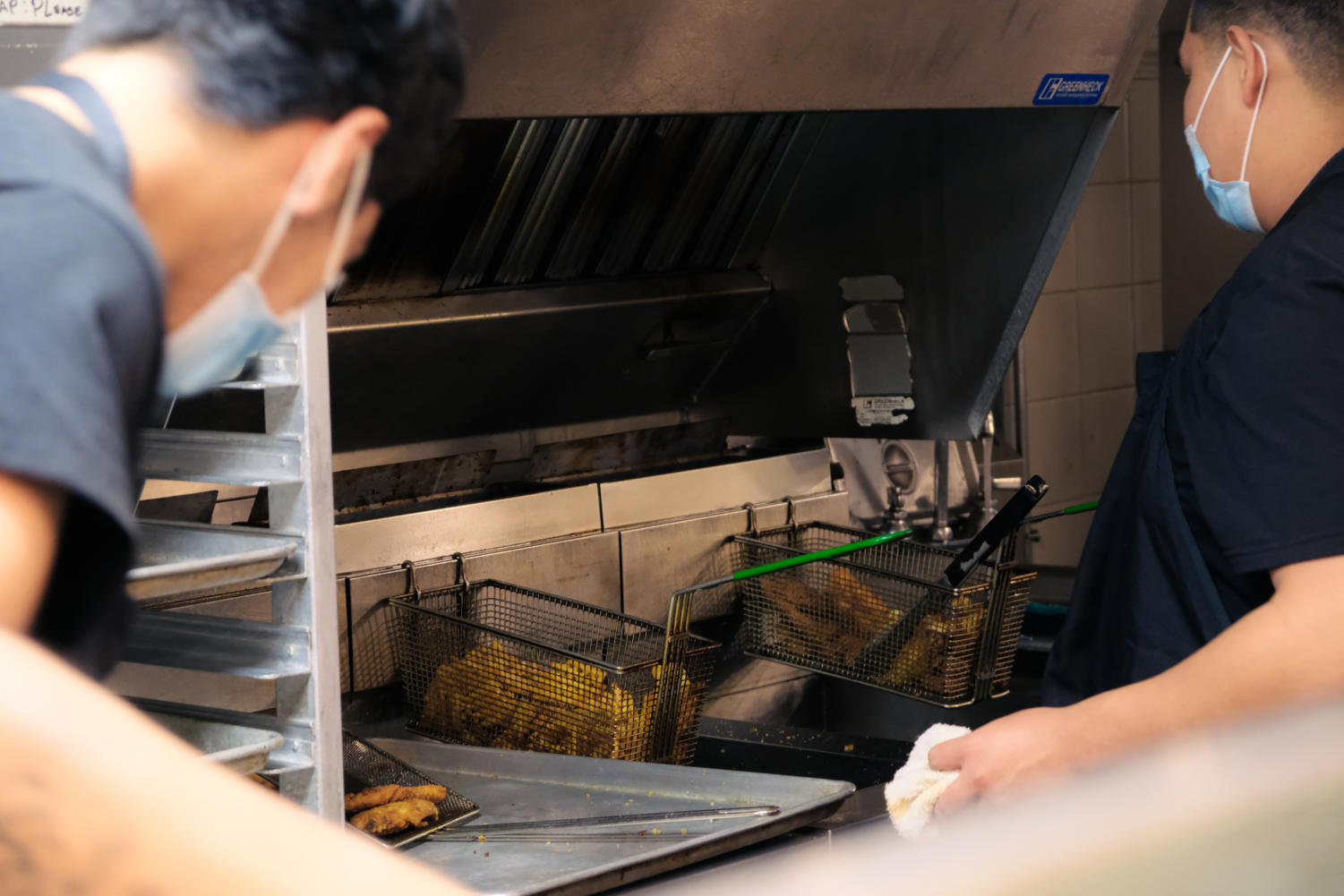 French fries and chicken wings in an oil fryer. Two staff members wearing masks watch.