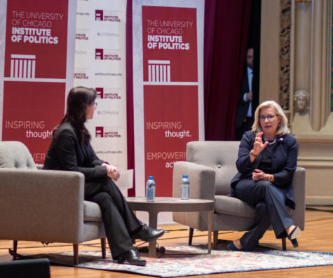 Liz Cheney, wearing a blue pantsuit, sits on a gray accent chair and gestures at the camera while Katherine Baicker, sitting profile to the camera, looks at her. A coffee table separates the pair.