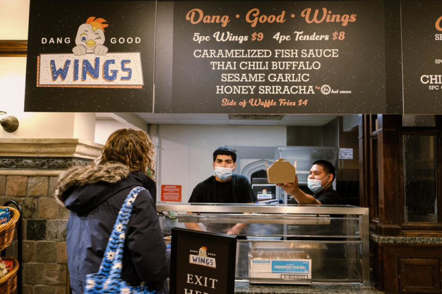 A+staff+member+at+Dang+Good+Wings+hands+over+a+brown+paper+box+to+a+customer+standing+at+the+counter.+Another+worker+stands+behind+the+counter.+The+Dang+Good+Wings+sign+advertises+5pc+wings+for+%249%3B+4pc+tenders+for+%248%3B+a+side+of+waffle+fries+for+%244%3B+and+caramelized+fish+sauce%2C+thai+chili+buffalo+sauce%2C+sesame+garlic+sauce%2C+and+honey+sriracha+sauce.