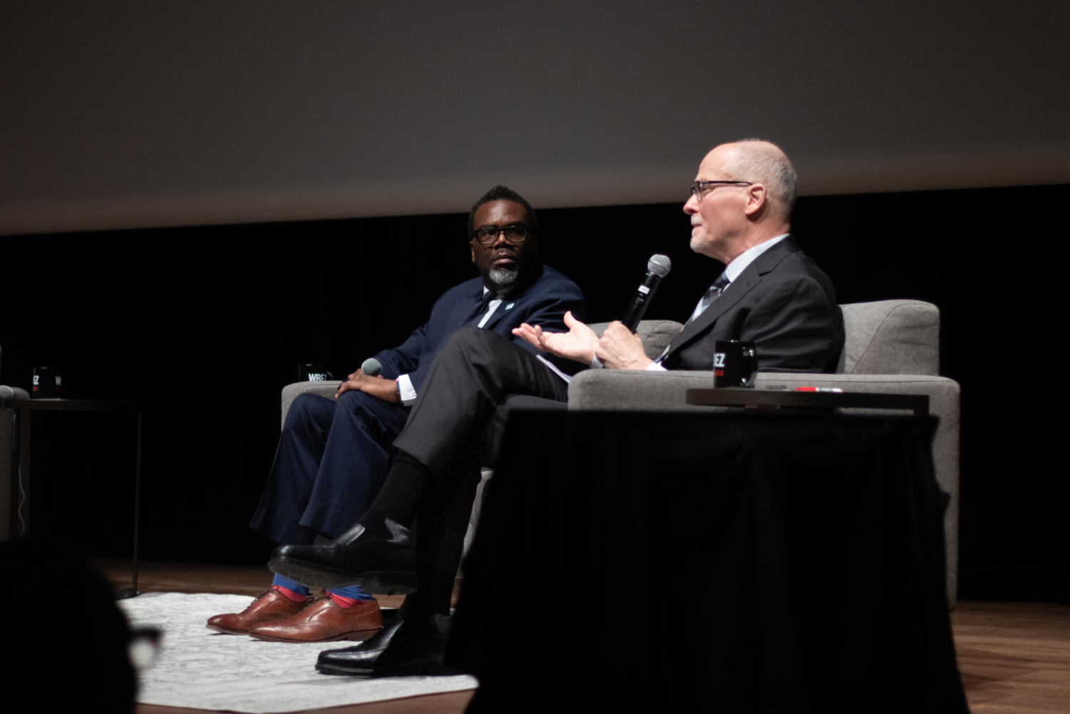 Brandon Johnson and Paul Vallas hold microphones while sitting in chairs. Vallas is speaking.